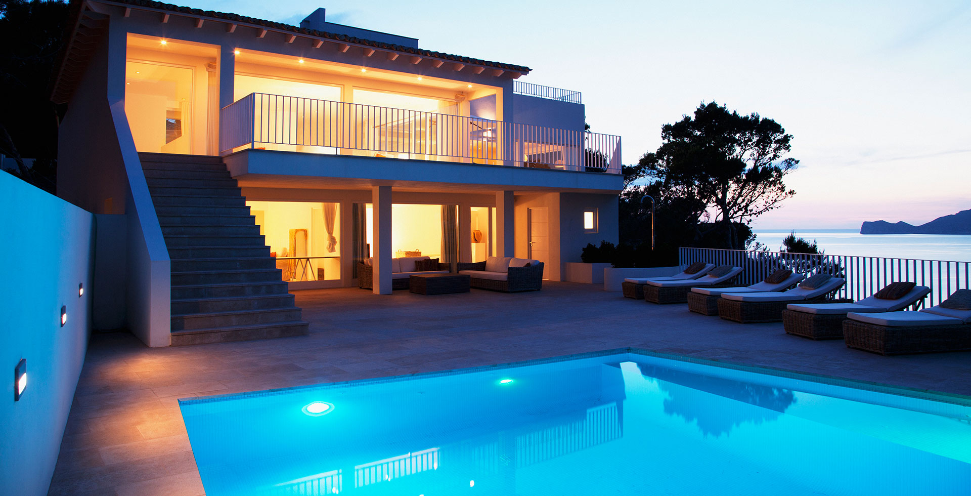 Exterior back view of a house with the interior lighting turned on. There is a swimming pool with swimming pool lighting in the foreground with six pool loungers in the background